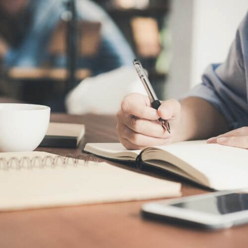 Sobre una mesa de madera, alguien escribe a mano en un cuaderno.