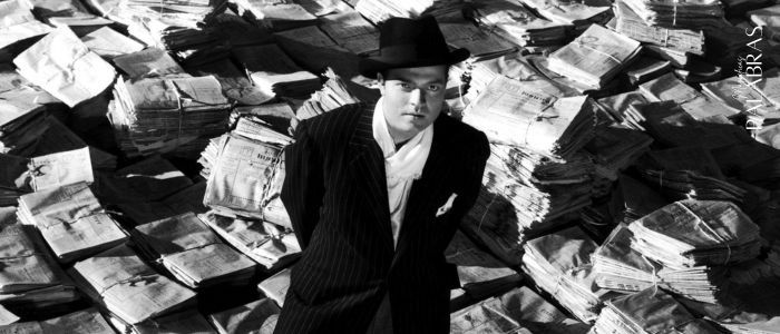 Imagen de la película Ciudadano Kane con Orson Welles sobre un montón de periódicos. Lleva abrigo y sombrero. Cómo escribir un folleto, Magníficas Palabras Copywriting.